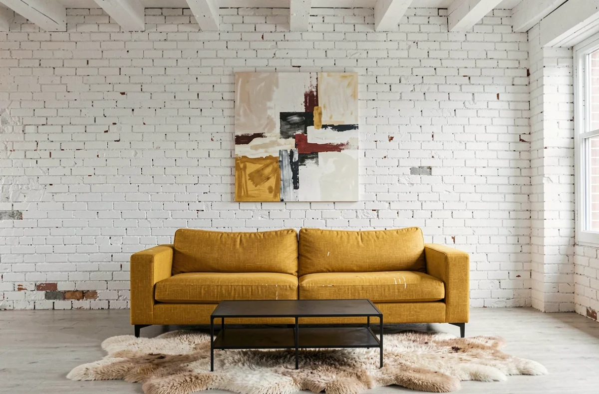 Loft contemporain avec murs en briques blanches et canapé jaune moutarde, tapis en fausse fourrure et œuvre d'art abstraite au mur